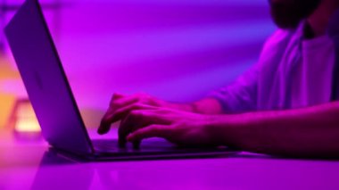 Close up, hands behind a laptop in neon light. Programming, video game, computer work in the evening