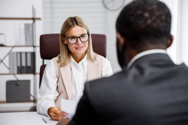 Hiring, interview. Middle-aged blond business woman conducting an interview in a bright office, an African American man. He looks at his resume and after reading it, shakes hands and smiles