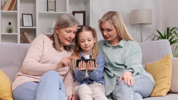白髪の女性の祖母 小さな孫娘とブロンドの母親は 母親の日にビデオを見てリビングルームに座っています かわいい女の子は彼女の家族に電話でプログラムを使用する方法を説明します — ストック動画