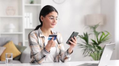 Masada oturan cep telefonu ve kredi kartıyla gülümseyen güzel Hintli esmer kız. Mutlu kadın mobil ödemeyi kullanarak online alışveriş yapıyor. E-bankacılık uygulama servisi