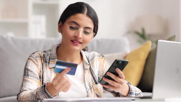 笑着年轻漂亮的印度黑发女孩拿着手机和信用卡坐在桌旁 快乐的女人用手机购物 电子银行应用软件服务 — 图库视频影像