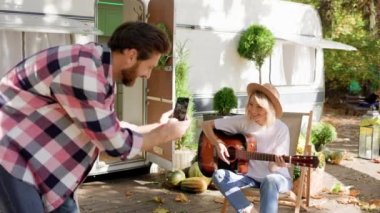 Komik ve neşeli genç bir çift. Kız gitar çalıyor, adam onu sosyal ağlar için bir seyahat karavanının arka planında fotoğraflıyor. Yaz mevsiminde hafta sonları. Doğada romantizm