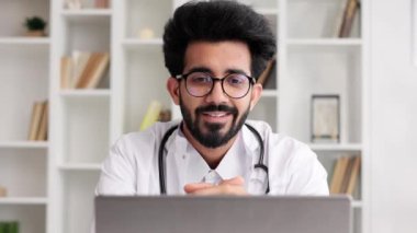 Beyaz önlüklü akıllı, dost canlısı Hintli klinisyen hastane ofisinde uzaktan hasta muayenesi için dizüstü bilgisayar kullanıyor. Teletıp. Bir doktorla çevrimiçi randevu.