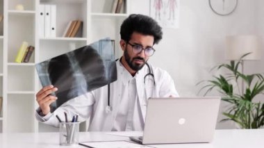 Beyaz önlüklü akıllı, dost canlısı Hintli klinisyen hastane ofisinde röntgenlere bakan hasta muayenesi için dizüstü bilgisayar kullanıyor. Teletıp. Bir doktorla çevrimiçi randevu.