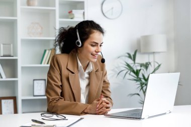 Young curly brunette woman wearing video call headset on laptop in home light office. Video conference, remote meeting. Programmer, remote work, work from home