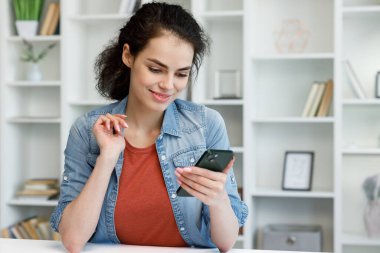 Young curly brunette sitting at the table uses a mobile phone. Surfs the Internet, communicates in instant messengers on his smartphone. Online communication, smartphone in human life