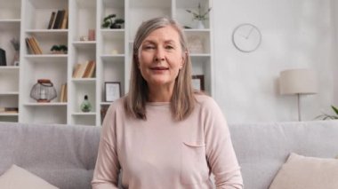 50 'li yaşlarda neşeli yaşlı bir kadın çevrimiçi görüşme sırasında evdeki kameraya bakıyor. Gülümseyen büyük anne kamerayla konuşuyor, görüntülü sohbet ediyor, internette sohbet ediyor, kanepede oturuyor, webcam izliyor.