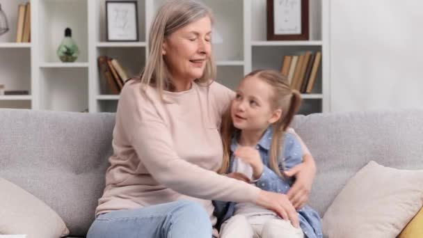 백발의 할머니와 손녀가 소파에 껴안고 집에서 즐겁게 시간을 보내고 세대를 — 비디오