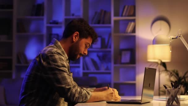 一个年轻的阿拉伯学生晚上在客厅的笔记本电脑上工作 一个留着胡子的男人在家里做It工作 敲键盘 在笔记本上写字 — 图库视频影像