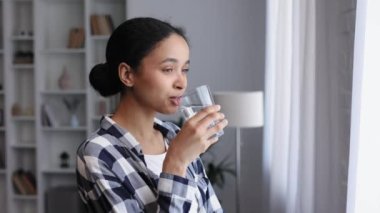 Genç bir Afrikalı Amerikalı kız oturma odasının penceresinin yanında duruyor ve yeni bir güne iyi bir alışkanlıkla başlıyor, bir bardak temiz su tutuyor ve onu içiyor, yakından bakıyor.