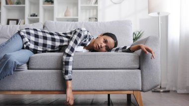 Yorgun Afrikalı Amerikalı kız kanepeye uzanmış uyuşuk hissediyor. Depresyonda, motivasyon eksikliği, üzüntü, can sıkıntısı. Yorgunluktan, stresten kanepede yığılıp kalıyor. Tükenmiş. Görüntüyü kapat