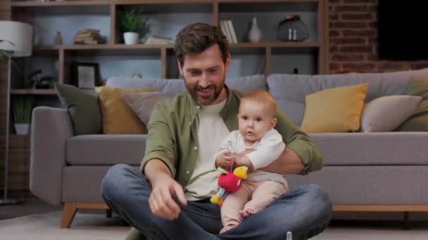 爸爸正在休产假 他抱着孩子坐在公寓的地毯上 用玩具干扰婴儿在智能手机上冲浪 — 图库视频影像