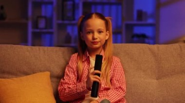 Neşeli kız akşamları kanepede oturuyor ve uzaktan kumandayla televizyonu açıyor. Kız en sevdiği çizgi film programı yayınlandığı için mutlu, ellerini kaldırıyor, izlemekten zevk alıyor.