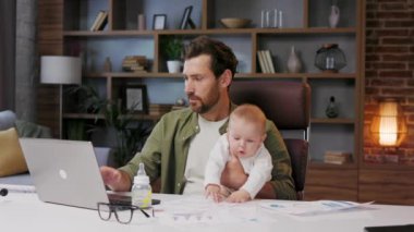 Father is on maternity leave, working in a home office at a laptop with a small child in his arms. Freelance businessman combines child care and work. Paternity Leave