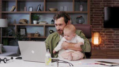 Portrait of a Father on maternity leave, in his home office with a small child in his arms. Dad at the table with a laptop and a child. Holiday to care for the child. Maternity leave for a man
