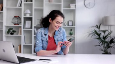 Young curly brunette sitting at the table uses a mobile phone. Surfs the Internet, communicates in instant messengers on his smartphone. Online communication, smartphone in human life