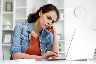 Young curly brunette, sitting stressed at the table and working on a laptop in a bright home office, apathy and depression from overwork at work. Burnout from the modern world. Programmer, student.