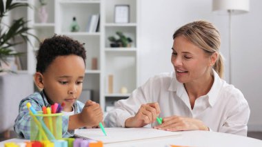 Adorable little african american boy with curly hair having fun at pediatric specialist appointment, happy child drawing with female therapist, exercises for children with autism clipart