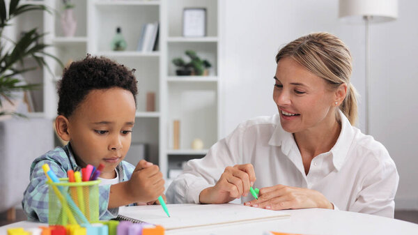 Adorable little african american boy with curly hair having fun at pediatric specialist appointment, happy child drawing with female therapist, exercises for children with autism