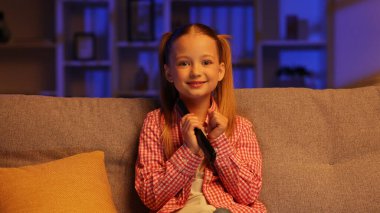 Neşeli kız akşamları kanepede oturuyor ve uzaktan kumandayla televizyonu açıyor. Kız en sevdiği çizgi film programı yayınlandığı için mutlu, ellerini kaldırıyor, izlemekten zevk alıyor.