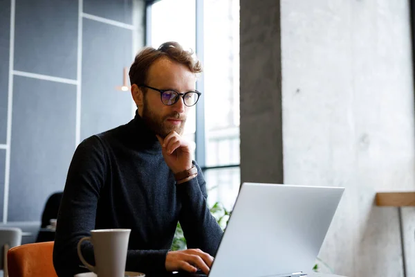 stock image A young red-haired guy, a programmer or entrepreneur in glasses, works in a stylish cafe behind a laptop. Freelancer works remotely. Online communication. Small business