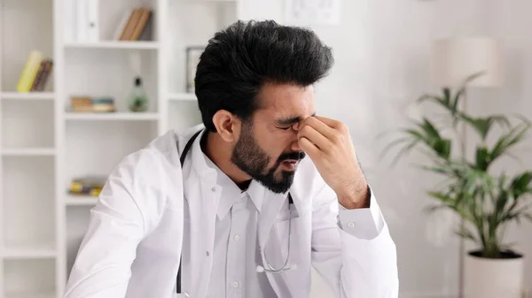 Stressed young male Indian doctor in white uniform with stethoscope looking at laptop screen, thinking about difficult solution to medical problem or feeling exhausted after hard day in hospital