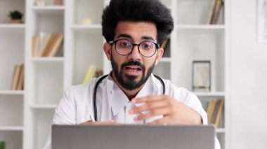 Beyaz önlüklü akıllı, dost canlısı Hintli klinisyen hastane ofisinde uzaktan hasta muayenesi için dizüstü bilgisayar kullanıyor. Teletıp. Bir doktorla çevrimiçi randevu.