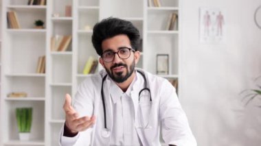 Beyaz önlüklü akıllı, dost canlısı Hintli klinisyen hastane ofisinde uzaktan hasta muayenesi için dizüstü bilgisayar kullanıyor. Teletıp. Bir doktorla çevrimiçi randevu. Kamera görünümü
