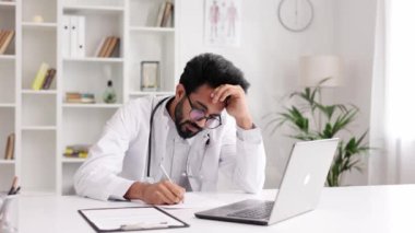 Stresli Hintli beyaz üniformalı genç erkek doktor dizüstü bilgisayara bakıyor, tıbbi probleme zor bir çözüm düşünüyor ya da hastanede geçirdiği yorucu bir günün ardından bitkin düşüyor.