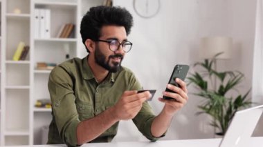 Elinde cep telefonu ve kredi kartı olan gülümseyen Hintli esmer çocuk masada oturuyor. Mutlu adam mobil ödemeyi kullanarak online alışveriş yapıyor. E-bankacılık uygulama servisi