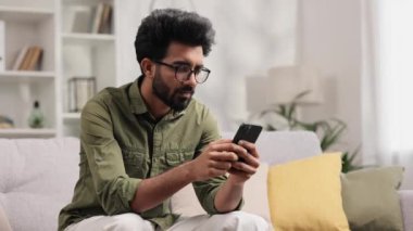 Kanepede oturup telefonuna bakan ve neden çalışmadığını anlamayan endişeli genç Hintli çocuk. Gözlüklü kızgın öğrenci gördüğü kötü habere inanmıyor.