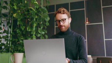 Genç kızıl saçlı bir adam, bir programcı ya da gözlüklü bir girişimci, bir dizüstü bilgisayarın arkasındaki şık bir kafede çalışıyor. Freelancer uzaktan çalışıyor. Çevrimiçi iletişim. Küçük işletme