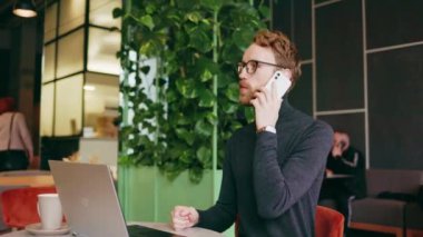 Genç kızıl saçlı bir adam, bir programcı ya da gözlüklü bir girişimci, şık bir kafede dizüstü bilgisayarda oturuyor ve telefonda konuşuyor. Freelancer uzaktan çalışıyor. Çevrimiçi iletişim. Küçük işletme
