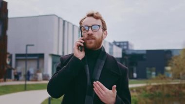 Genç, kızıl saçlı bir adam, gözlüklü, bir iş merkezinin arka planında, şık bir ceketle. Telefonda güvenli bir şekilde konuşuyor, hareket halindeyken sorunları çözüyor. Başarılı bir gencin portresi.