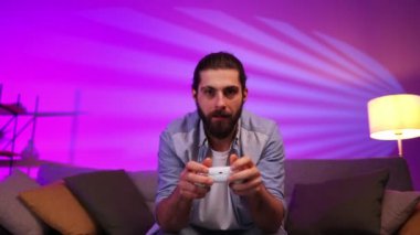 Neşeli genç adam TV 'nin önünde kanepede otururken konsolda video oyunları oynuyor. Oyuncu evde birlikte eğleniyor. Neon ışıklı oda