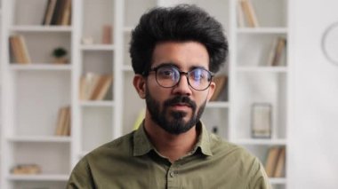 Çekici, kendine güvenen, dişlek gülüşlü, modern oturma odasında kameraya bakan Hintli adamın muhteşem portresi. İş adamı ev ofisinde şık bir görünüm takınıyor.