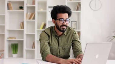 Çekici, kendine güvenen, dişlek gülüşlü, modern oturma odasında kameraya bakan Hintli adamın muhteşem portresi. İş adamı ev ofisinde şık bir görünüm takınıyor.