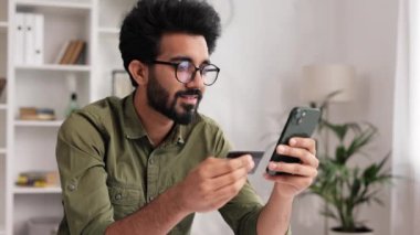 Elinde cep telefonu ve kredi kartı olan gülümseyen Hintli esmer çocuk masada oturuyor. Mutlu adam mobil ödemeyi kullanarak online alışveriş yapıyor. E-bankacılık uygulama servisi
