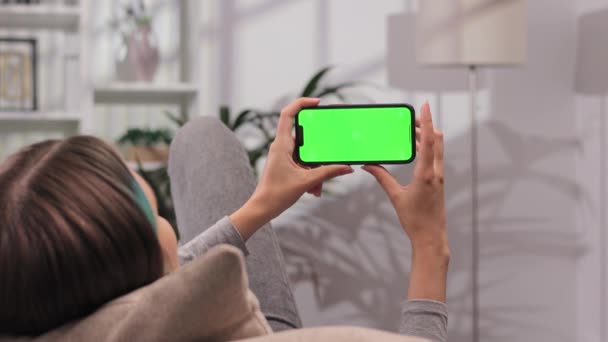 明るい部屋で緑色のスクリーンモックアップで横の位置にスマートフォンを使用してソファーに横たわる少女のクローズアップショット ビデオを見ながらさまざまなジェスチャーを使用 — ストック動画