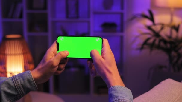 夜の部屋で緑色のスクリーンモックアップで横の位置でスマートフォンを使用してソファーに座っている男のクローズアップショット ゲームに2本の指を使用します — ストック動画