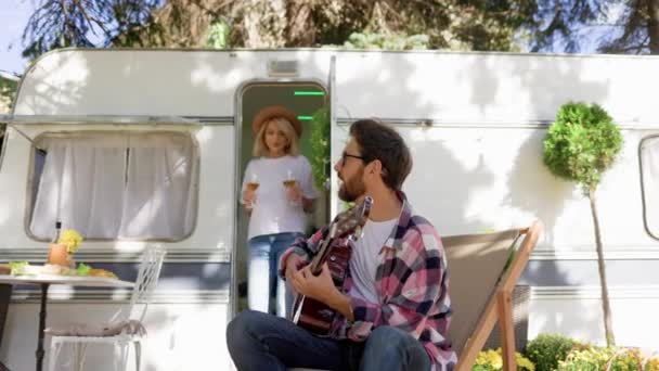 那家伙在旅行拖车前弹奏吉他 这个女孩提供了一杯饮料 在城外休息 快乐的人享受 夏天的周末 浪漫的本质 — 图库视频影像