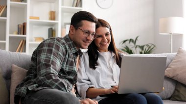 Oturma odasındaki kanepede dizüstü bilgisayarlı genç bir çift oturuyor, alışveriş yapıyorlar, tatil paketleri seçiyorlar. Bir erkek ve bir kadın uzun zamandır beklenen alışverişe sevinir, birlikte mutlu olurlar.