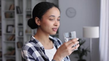 Genç bir Afrikalı Amerikalı kız oturma odasının penceresinin yanında duruyor ve yeni bir güne iyi bir alışkanlıkla başlıyor, bir bardak temiz su tutuyor ve onu içiyor, yakından bakıyor.