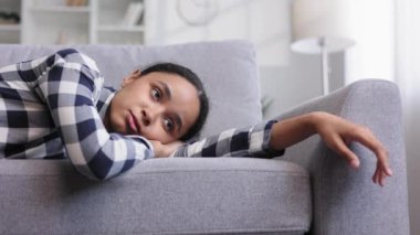 Yorgun Afrikalı Amerikalı kız kanepeye uzanmış uyuşuk hissediyor. Depresyonda, motivasyon eksikliği, üzüntü, can sıkıntısı. Yorgunluktan, stresten kanepede yığılıp kalıyor. Tükenmiş. Görüntüyü kapat