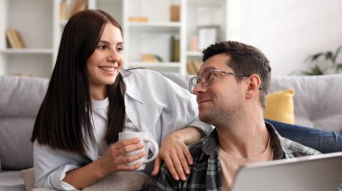 Neşeli genç çift, dizüstü bilgisayar ve kahveyle birlikte oturma odasında dinleniyor, mutlu genç çift sıcak içecekler içiyor, bilgisayar kullanıyor ve alışveriş yapıyor, yakın çekim yapıyor.
