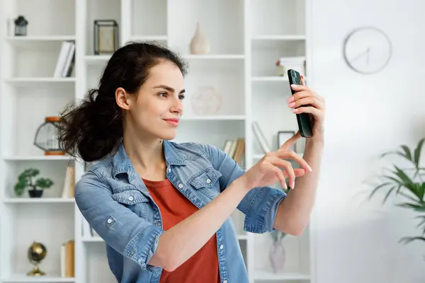 stock image Young curly brunette sitting at the table uses a mobile phone. A vidoe call communicates in instant messengers on your smartphone. Online communication, smartphone in human life