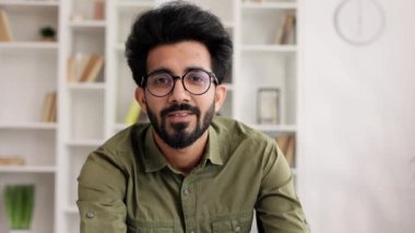 Çekici, kendine güvenen, dişlek gülüşlü, modern oturma odasında kameraya bakan Hintli adamın muhteşem portresi. İş adamı ev ofisinde şık bir görünüm takınıyor.