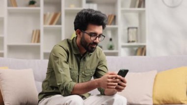 Siyah gözlüklü memnun Hintli adam e-postalarını kontrol ediyor ve iyi haberlerle mutlu. Kazanan genç adam zaferi kutluyor koltukta oturuyor, ellerini yukarı kaldırıyor.