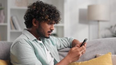 Kanepede oturup telefonuna bakan ve neden çalışmadığını anlamayan endişeli genç Hintli çocuk. Gözlüklü kızgın öğrenci gördüğü kötü habere inanmıyor.