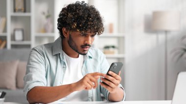 Kanepede oturup telefonuna bakan ve neden çalışmadığını anlamayan endişeli genç Hintli çocuk. Gözlüklü kızgın öğrenci gördüğü kötü habere inanmıyor.